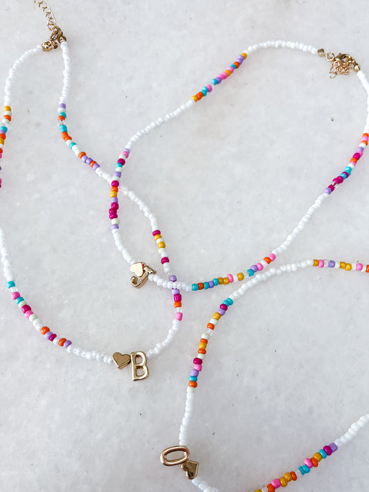 The Colorful Beaded Initial Necklace by Tiny Tot Bundles features three necklaces with colorful and white beads, each adorned with a gold heart charm and either the letter B, O, or the number 1, all displayed on a white background.