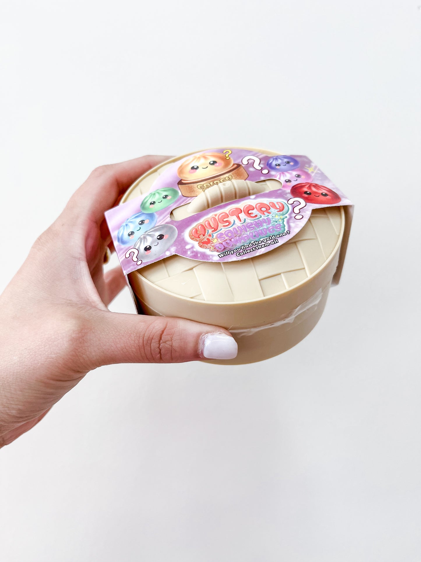 A hand with white nail polish holds a beige container with a colorful label featuring cute cartoon animals and "MYSTERY" in bold, indicating the Mystery Metallic Dumpling collectible toy from Prime Opticals.