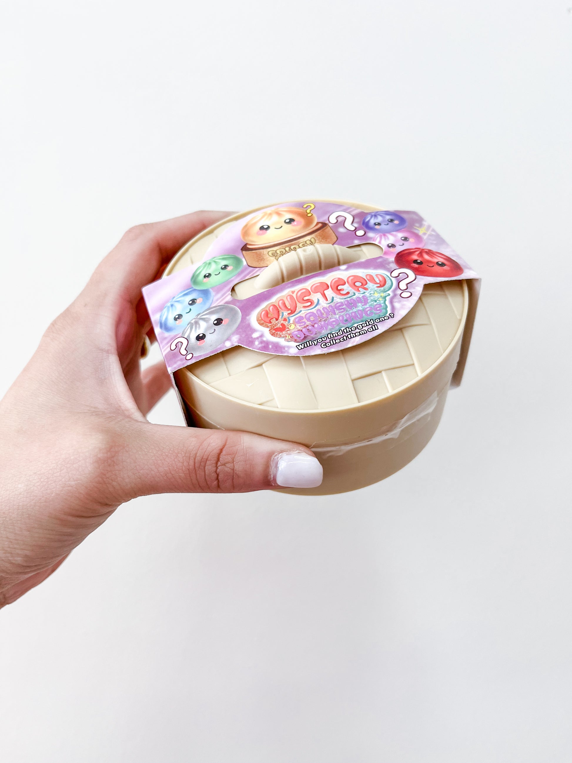 A hand with white nail polish holds a beige container with a colorful label featuring cute cartoon animals and "MYSTERY" in bold, indicating the Mystery Metallic Dumpling collectible toy from Prime Opticals.