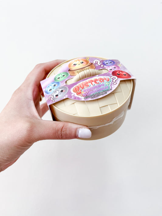 A hand with white nail polish holds a beige container with a colorful label featuring cute cartoon animals and "MYSTERY" in bold, indicating the Mystery Metallic Dumpling collectible toy from Prime Opticals.