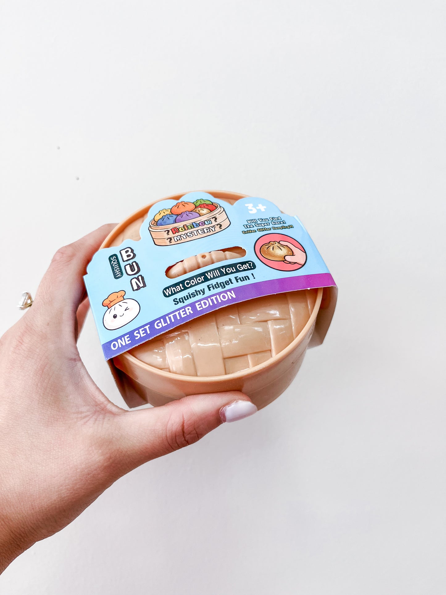 A hand holds a round container labeled Glitter MYSTERY Dumpling by Prime Opticals, featuring a cartoon dumpling and the text "What color will you get? Squishy Fidget Fun! One Set Glitter Edition" against a plain white background.