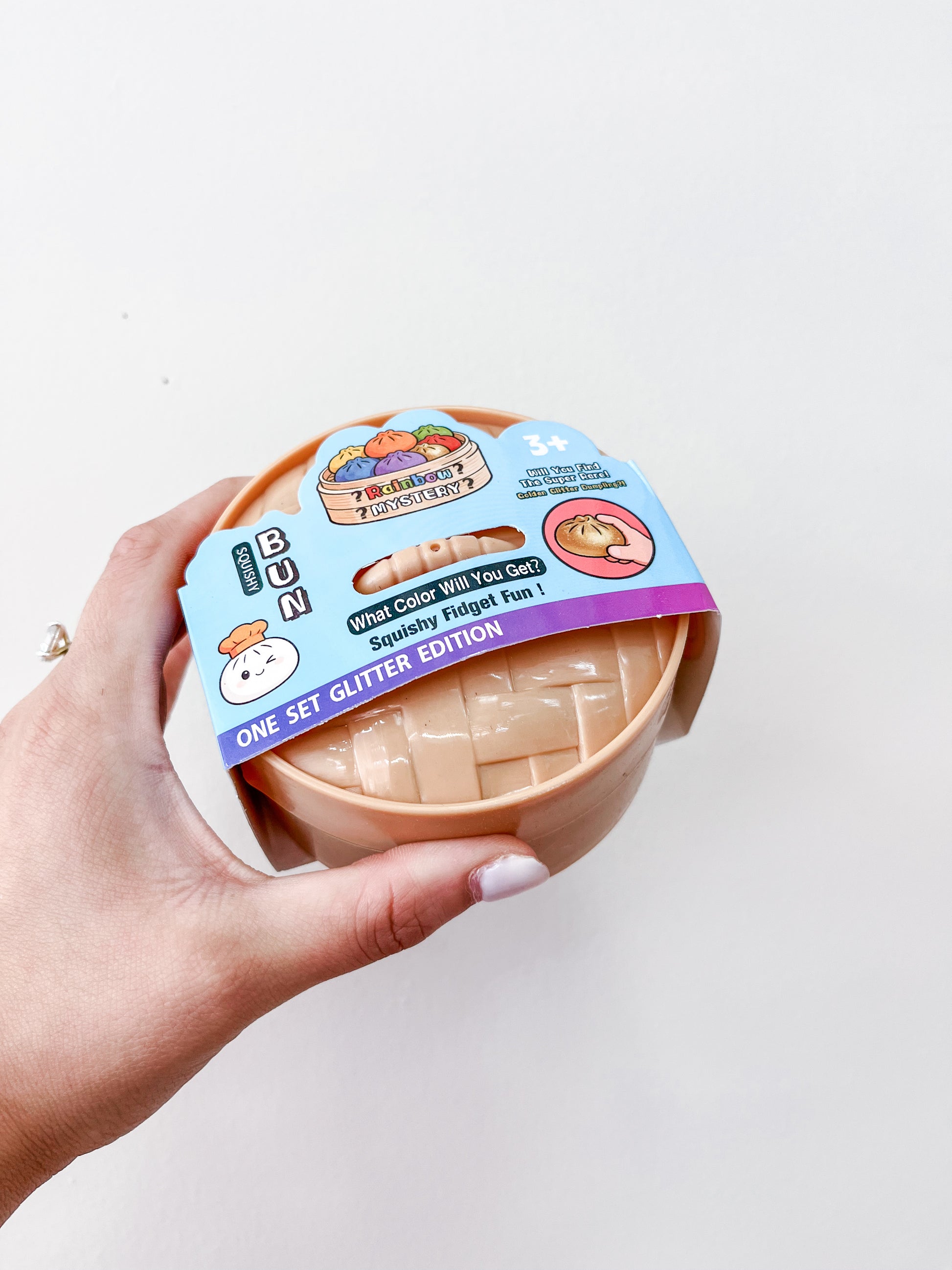 A hand holds a round container labeled Glitter MYSTERY Dumpling by Prime Opticals, featuring a cartoon dumpling and the text "What color will you get? Squishy Fidget Fun! One Set Glitter Edition" against a plain white background.
