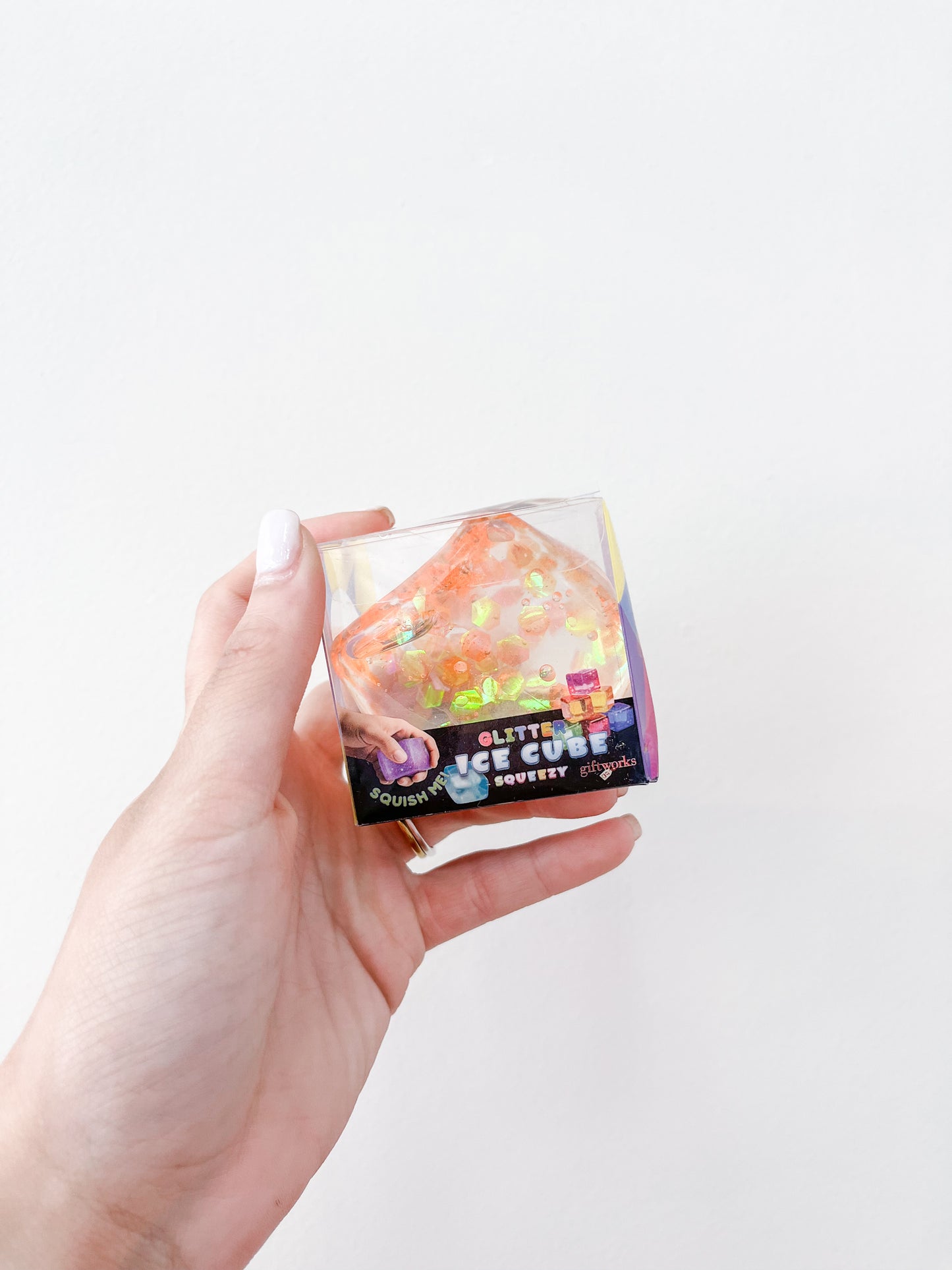 A hand with light-colored nail polish holds a small clear box containing a glittery orange triangular squishy toy labeled "Confetti Glitter Ice Cubes" by Prime Opticals against a plain white background.