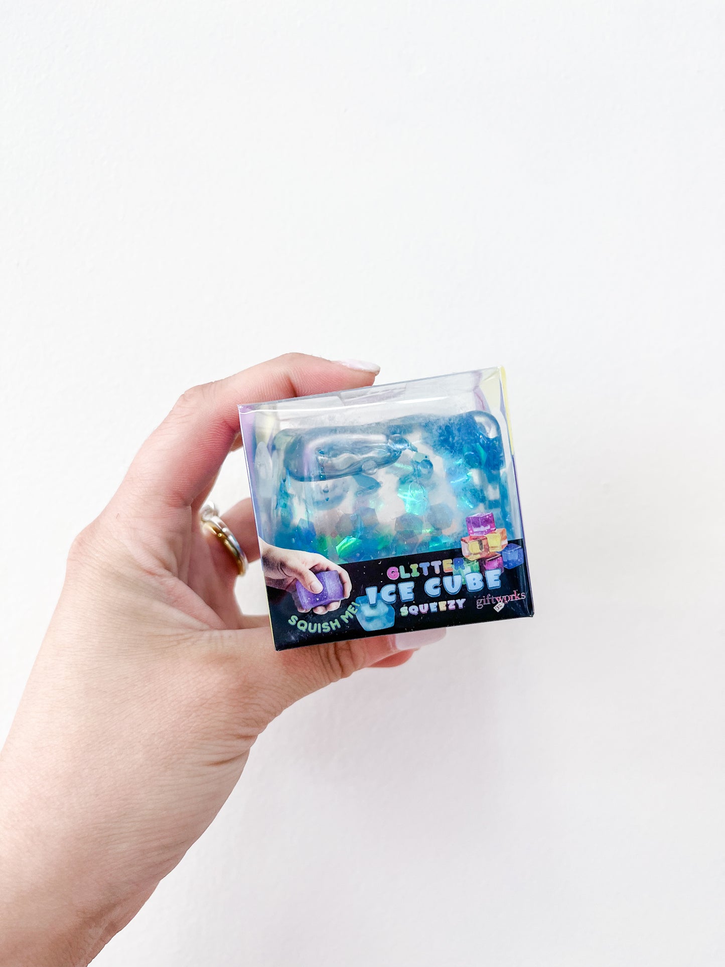 A hand holds a small clear box of Prime Opticals Confetti Glitter Ice Cubes, a blue squishy toy in colorful packaging with graphics, set against a plain white background.
