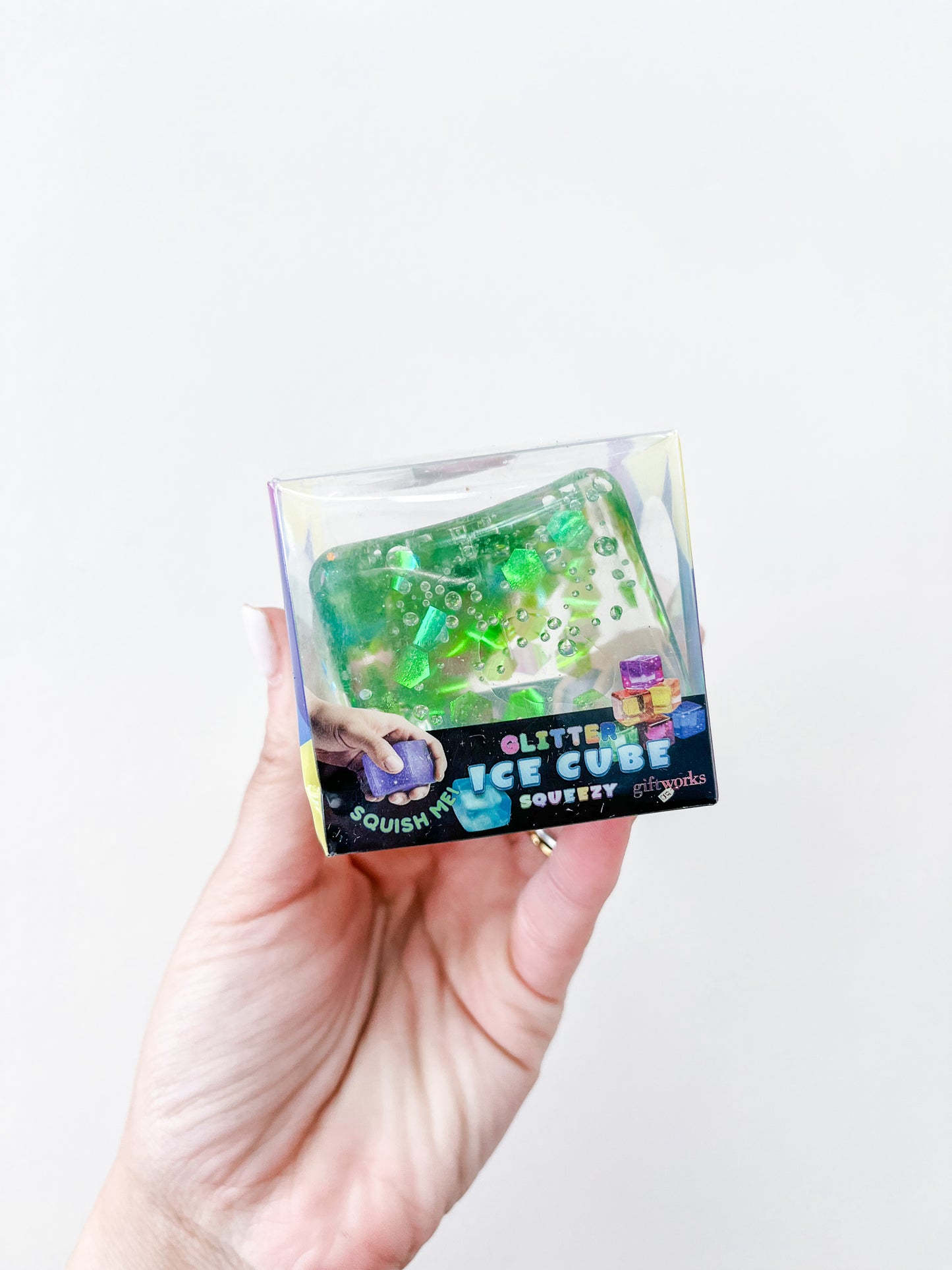 A hand holds a small, clear box with a green glittery ice cube squishy toy filled with beads. The packaging reads "Confetti Glitter Ice Cubes" by Prime Opticals and features colorful graphics.