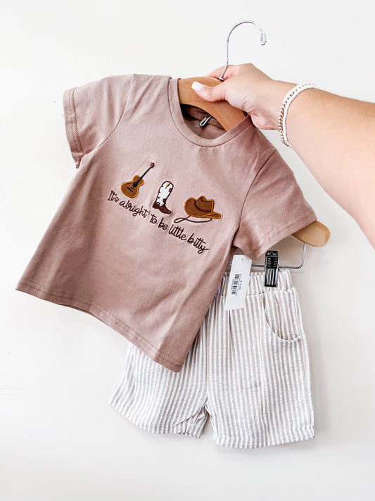 A hand holds up the Tiny Tot Bundles Itty Bitty Set: a brown t-shirt with guitar, boot, and hat graphics and the phrase "It's alright to be a little bitty," paired with light striped shorts.