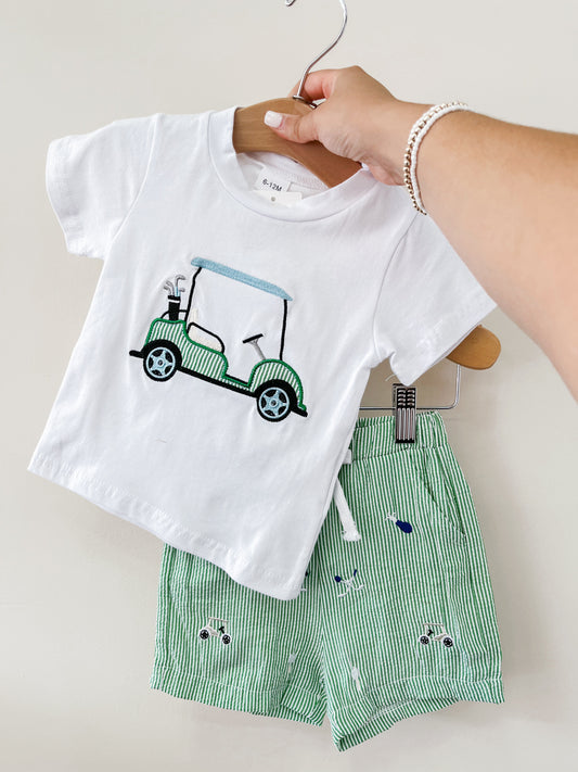 A hand holds up the Tiny Tot Bundles Golf Embroidered Set: a white t-shirt with a green golf cart graphic and green-and-white striped shorts featuring small blue and green embroidered cars.
