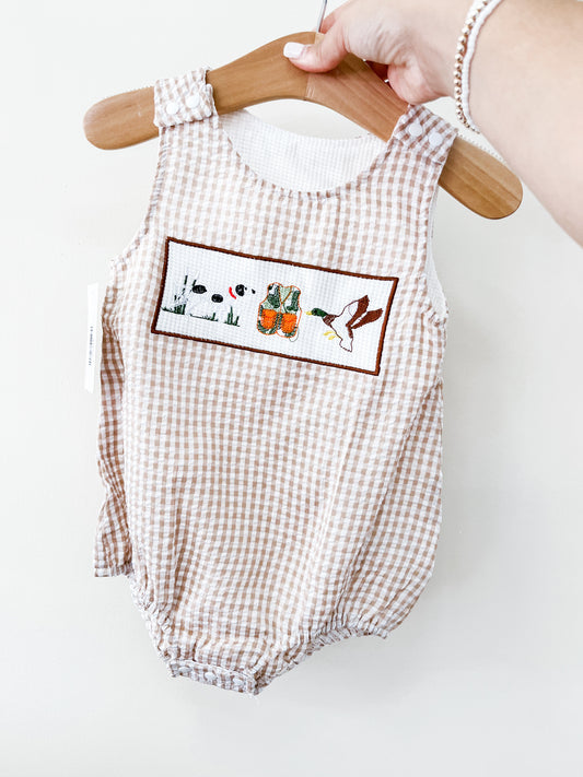A hand holds the Tiny Tot Bundles Mallard Duck Gingham Romper on a wooden hanger. This light brown and white romper features a chest patch embroidered with ducks, a dog, and reeds.