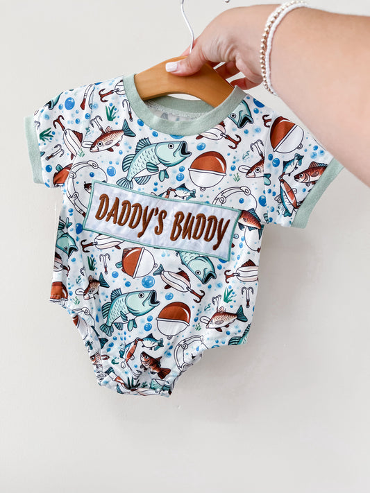 A hand holds the Tiny Tot Bundles Dad's Buddy Fishing Romper on a wooden hanger. The romper features fishing-themed designs with fish, lures, bobbers, and “DAD'S BUDDY” printed on the front.
