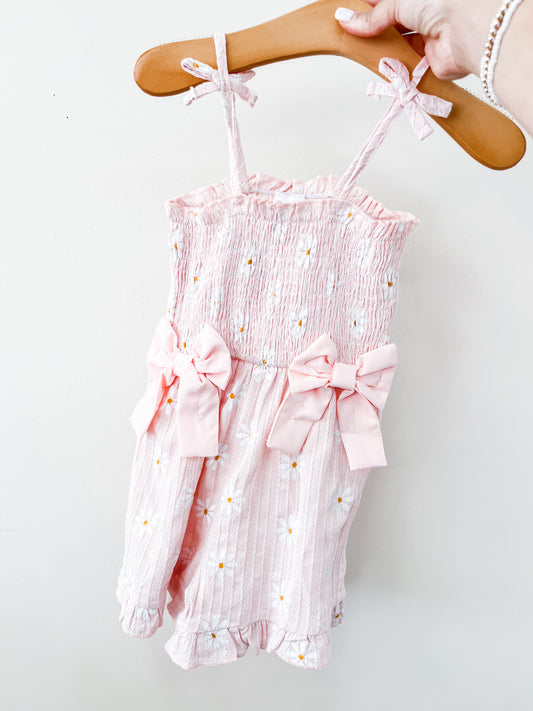 A hand holds the Tiny Tot Bundles Dainty Pink Scrunch Romper, a pink baby outfit with small white and yellow floral details, two large pink bows on the front, and ruffled straps and leg openings.