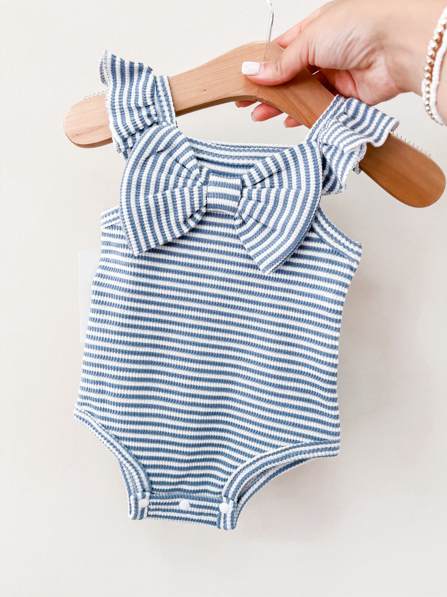 A hand holds the Tiny Tot Bundles Summer Blue Stripe Bow Romper on a wooden hanger, featuring ruffled straps and a large chest bow, against a plain light background.