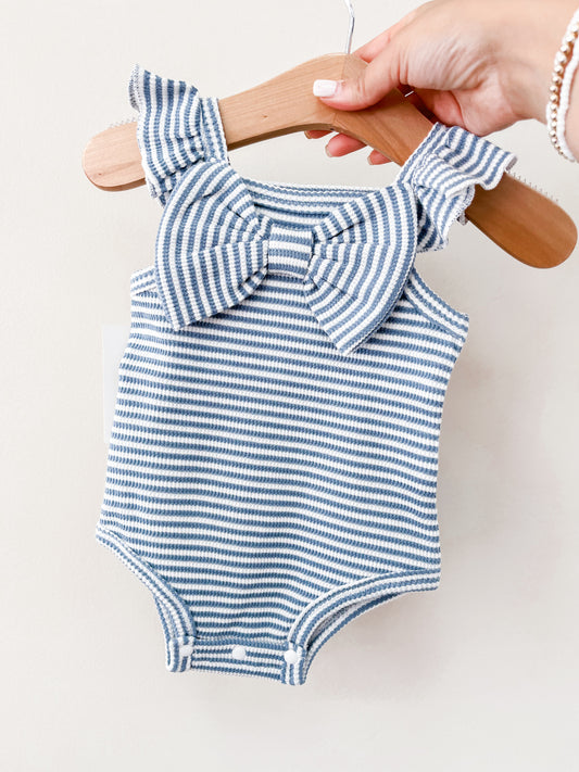 A hand holds the Tiny Tot Bundles Summer Blue Stripe Bow Romper on a wooden hanger, featuring ruffled straps and a large chest bow, against a plain light background.