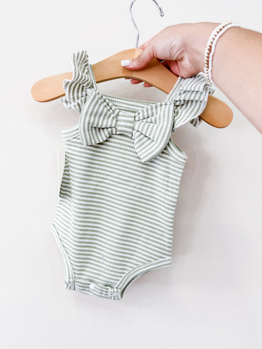 A hand holds the Tiny Tot Bundles Summer Green Stripe Bow Romper, featuring ruffled straps and a large chest bow, displayed on a wooden hanger against a plain white background.