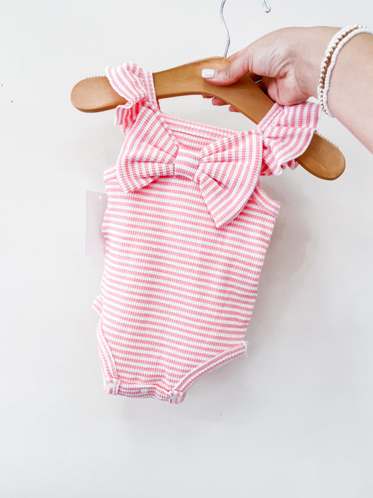 A hand holds the Tiny Tot Bundles Summer Pink Stripe Bow Romper—a baby outfit with pink and white stripes, ruffled shoulder straps, and a large front bow—on a wooden hanger against a plain white background.