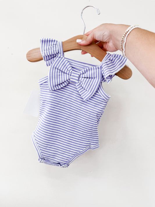 A hand holds the Tiny Tot Bundles Summer Purple Stripe Bow Romper, a sleeveless baby outfit with purple and white stripes, ruffled shoulders, and a front bow. The background is a plain white wall.