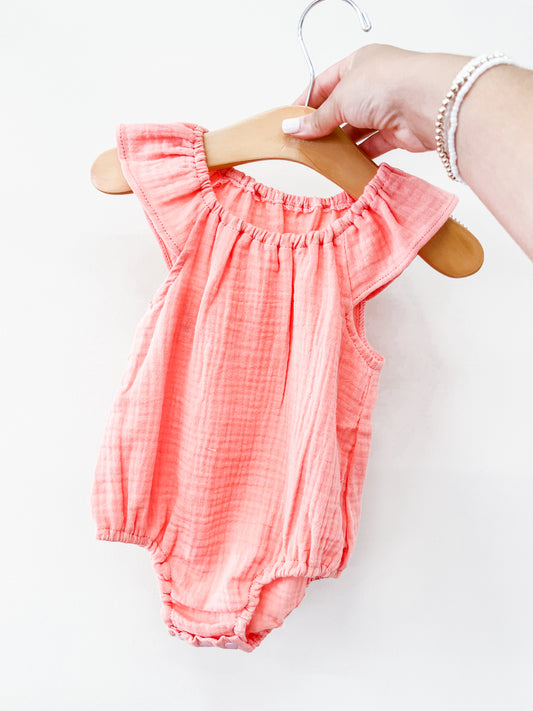 A hand holds the Tiny Tot Bundles Pink Summer Linen Romper, featuring ruffled sleeves and gathered edges, displayed on a wooden hanger against a plain white background.