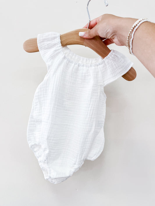 A hand holds a Tiny Tot Bundles White Summer Linen Romper, made of lightweight, textured fabric, displayed on a wooden hanger against a plain white background.