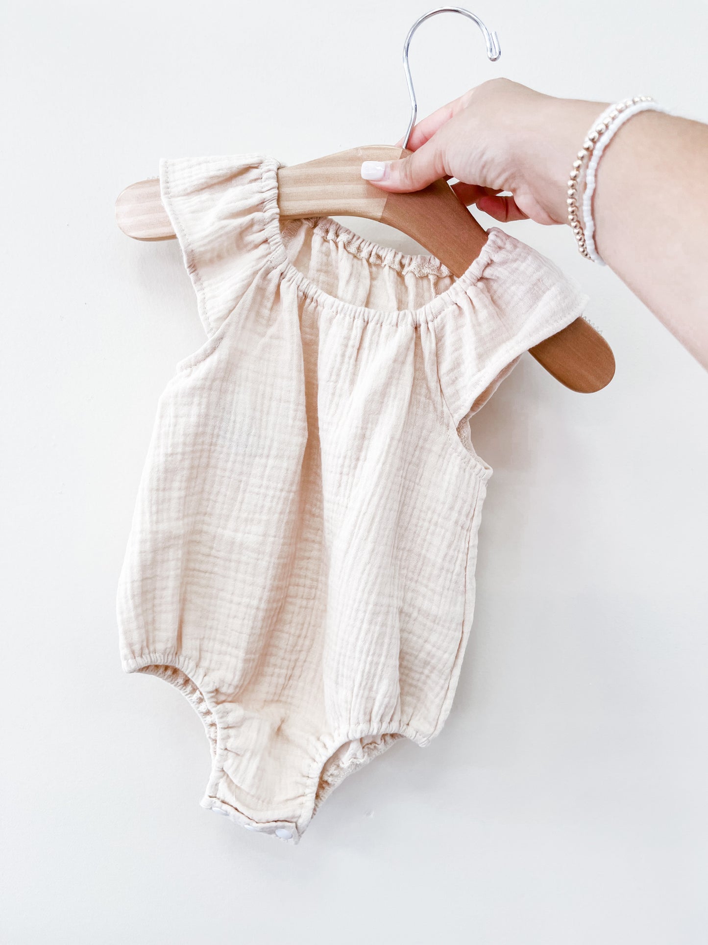 A hand holds the Tiny Tot Bundles Beige Summer Linen Romper, a sleeveless baby outfit with ruffled shoulder details, displayed on a wooden hanger against a plain white background.