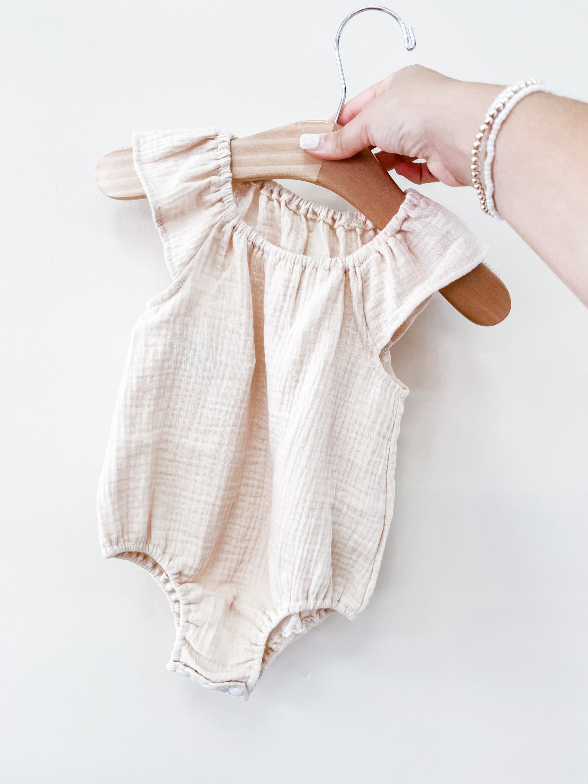 A hand holds the Tiny Tot Bundles Beige Summer Linen Romper, a sleeveless baby outfit with ruffled shoulder details, displayed on a wooden hanger against a plain white background.