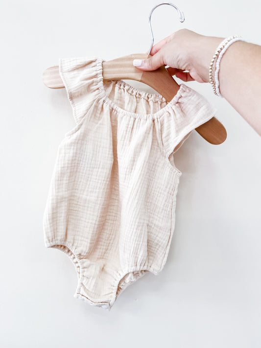 A hand holds the Tiny Tot Bundles Beige Summer Linen Romper, a sleeveless baby outfit with ruffled shoulder details, displayed on a wooden hanger against a plain white background.