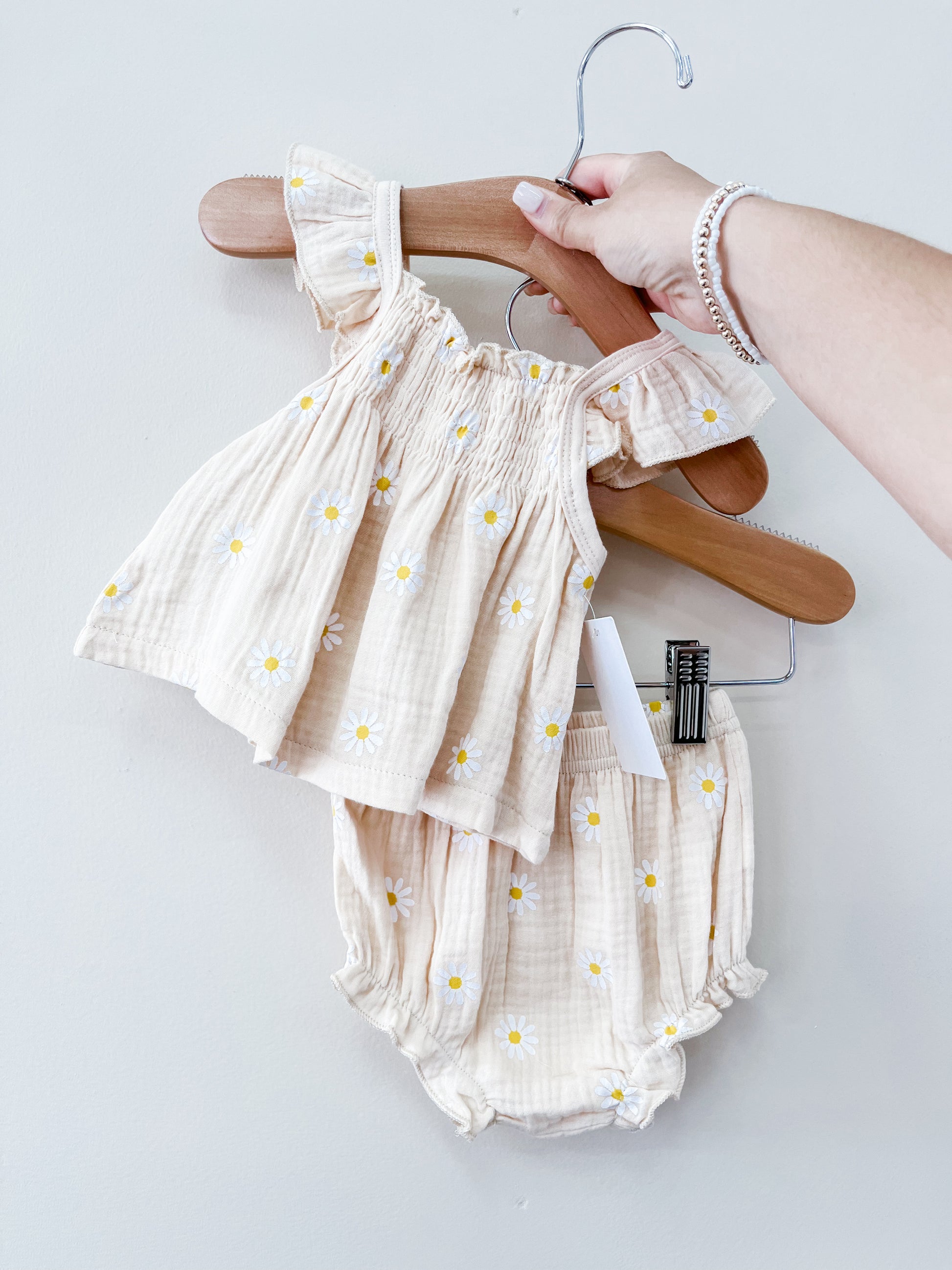 A hand holds the Tiny Tot Bundles Beige Daisy Set—matching baby top with ruffled sleeves and daisy prints, and coordinating bloomers—against a plain light background.