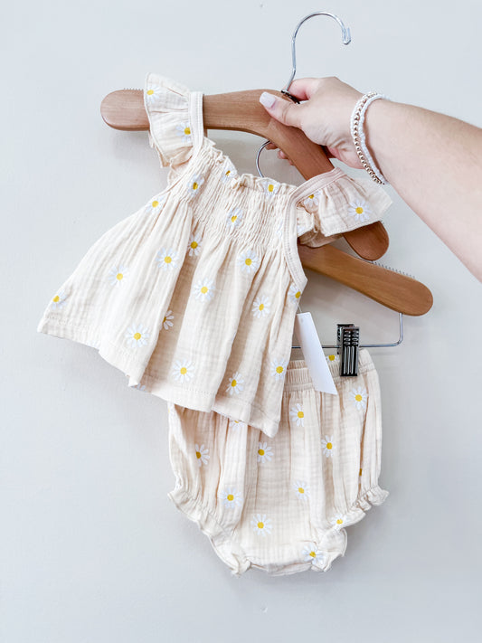 A hand holds the Tiny Tot Bundles Beige Daisy Set—matching baby top with ruffled sleeves and daisy prints, and coordinating bloomers—against a plain light background.