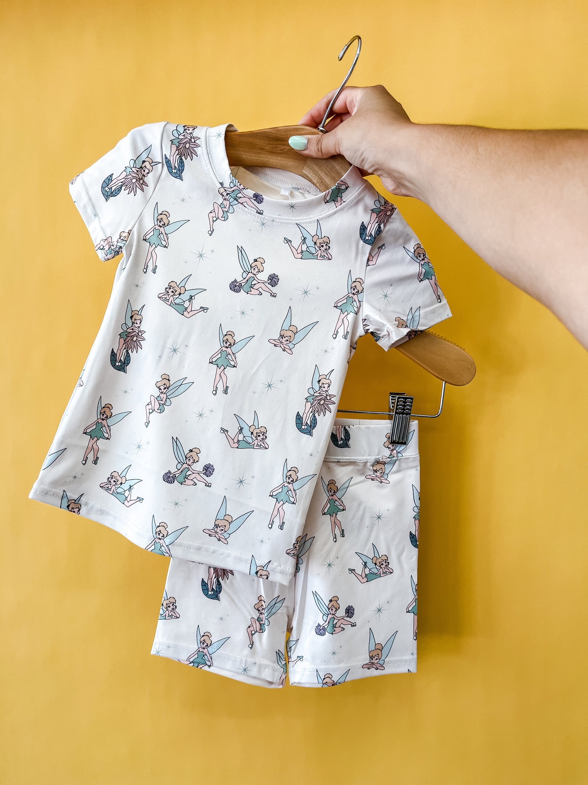 A hand holds a clothes hanger displaying the Tiny Tot Bundles Pixie Dust Lounge Set—a matching children’s T-shirt and shorts set adorned with fairy prints—against a yellow background.