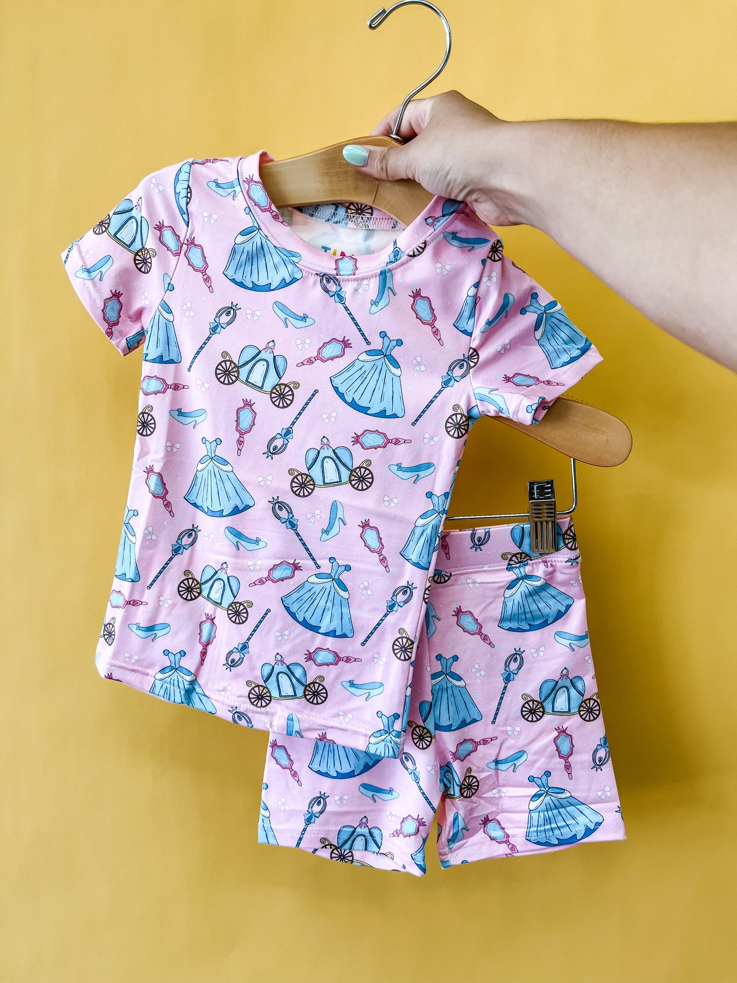 A hand holds the Tiny Tot Bundles Royal Ball Lounge Set, a pink children’s pajama set adorned with blue and purple princess-themed prints of dresses, shoes, mirrors, and carriages, against a solid yellow background.