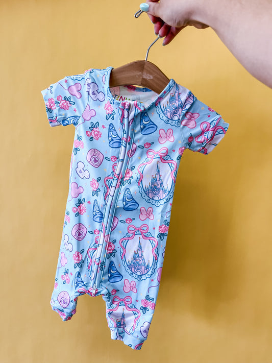 A hand holds up the Castle Charm Short Zippy by Tiny Tot Bundles on a hanger. The romper features a light blue color with pink and blue prints of castles, bows, and Mickey Mouse shapes, set against a yellow background.