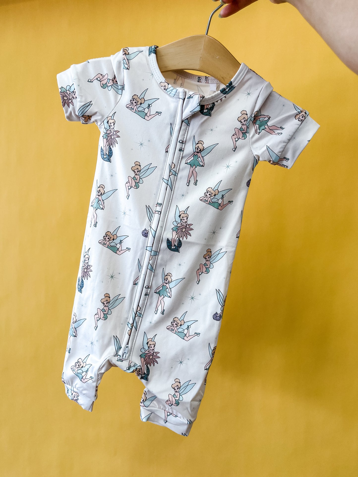 A hand holds the Tiny Tot Bundles Pixie Dust Short Zippy on a wooden hanger. The romper is white with a pattern of cartoon fairies and blue stars, set against a yellow background.