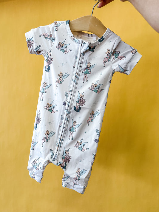 A hand holds the Tiny Tot Bundles Pixie Dust Short Zippy on a wooden hanger. The romper is white with a pattern of cartoon fairies and blue stars, set against a yellow background.