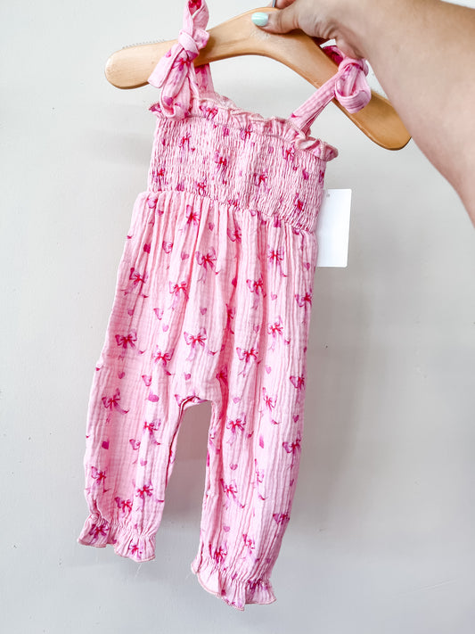 A hand holds up the Tiny Tot Bundles Blushing Bow Jumpsuit—a pink baby romper featuring shoulder ties, a ruffled top, and pink bow patterns—displayed on a wooden hanger against a plain white background.
