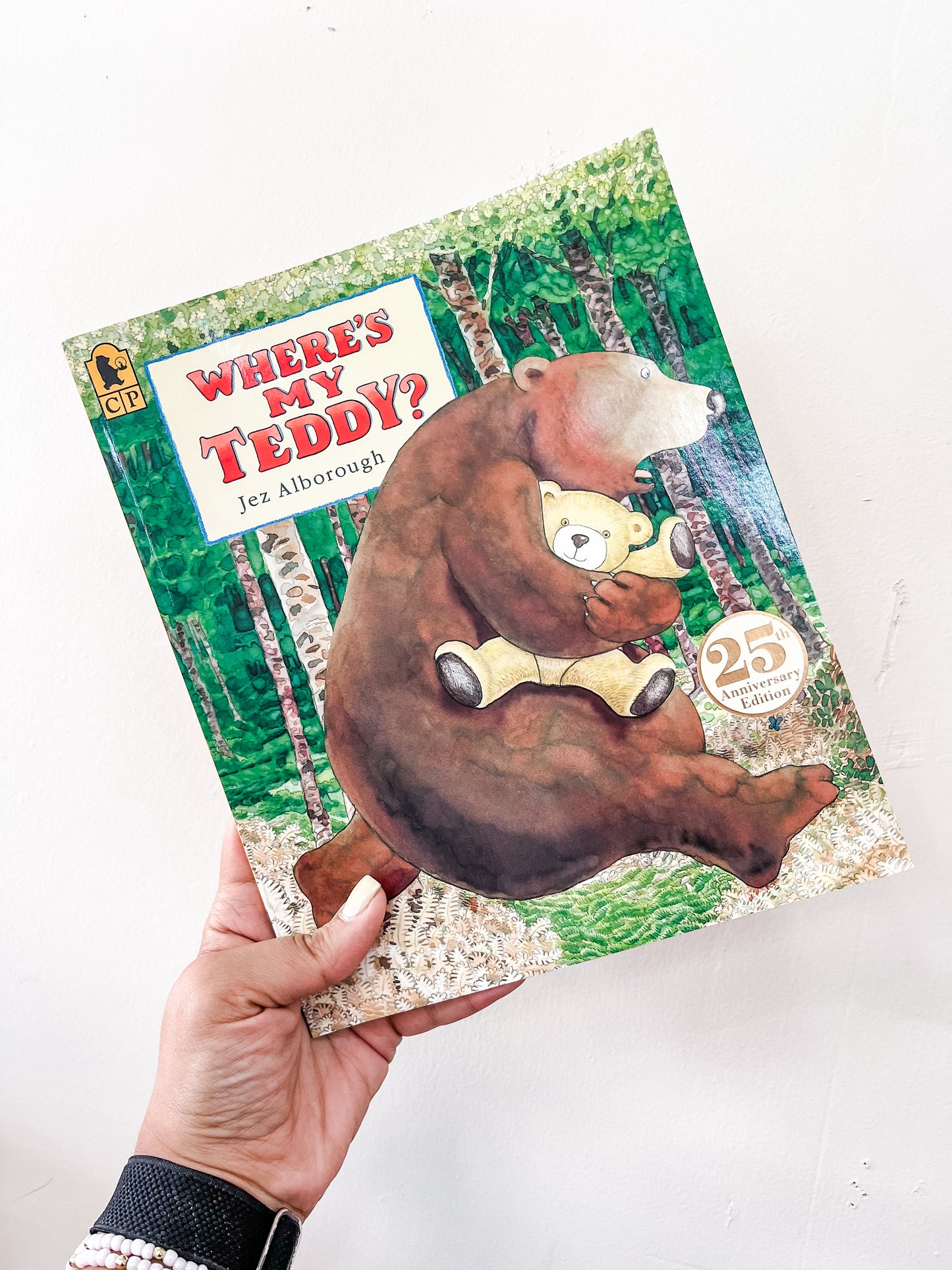 A hand holds the Tiny Tot Bundles Where's My Teddy Book, which displays a bear hugging a teddy bear in a forest and features a 25th Anniversary badge on the cover.