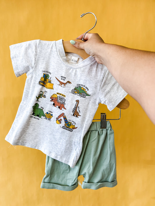 A hand holds a hanger displaying the Tiny Tot Bundles "Dino Construction Crew Set," which includes a white T-shirt with colorful cartoon vehicles and dinosaurs paired with light green shorts, all set against a yellow background.