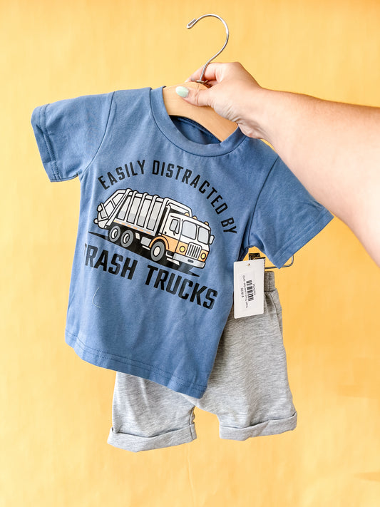 A hand holds the Tiny Tot Bundles "Trash Truck Lover Set," featuring a blue toddler t-shirt with a garbage truck graphic and the phrase “Easily Distracted by Trash Trucks,” paired with gray shorts, displayed on a hanger against a yellow background.