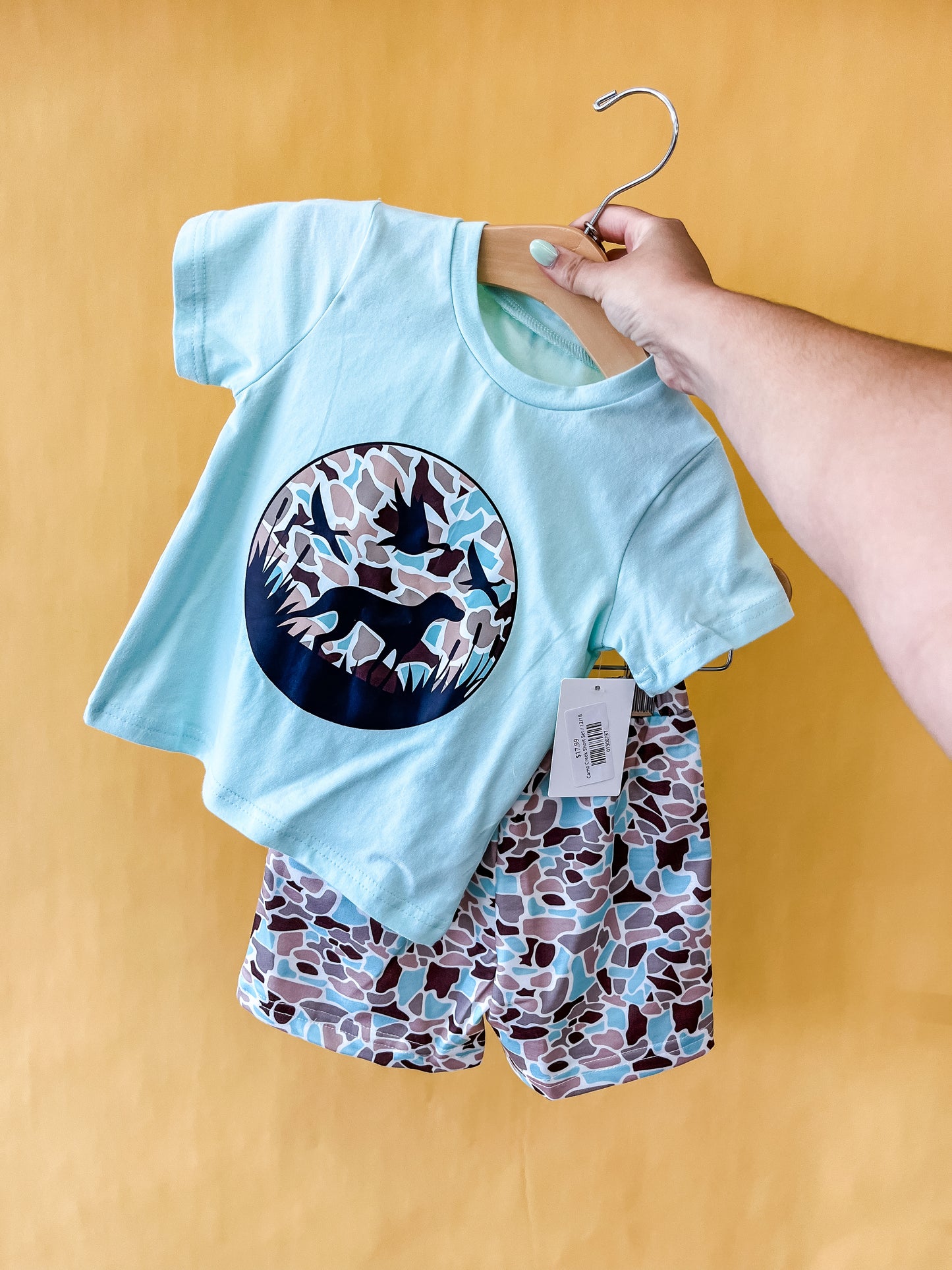 A hand displays the Camo Creek Short Set by Tiny Tot Bundles—a light blue T-shirt featuring a dinosaur graphic paired with matching camo-print shorts—against a yellow background.