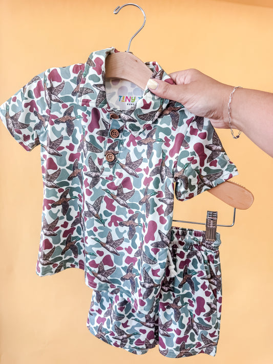 A hand holds a hanger displaying the Tiny Tot Bundles Mallard Marsh Collared Set—an adorable toddler’s matching camo shirt and shorts ensemble with a star pattern and wooden buttons—all set against a solid yellow background.