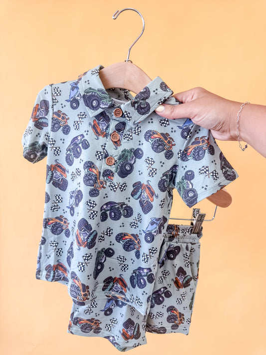 A hand holds up the Monster Mash Collared Set by Tiny Tot Bundles, featuring a collared matching shirt and shorts with a playful monster truck pattern, displayed on a wooden hanger against a peach-colored background.