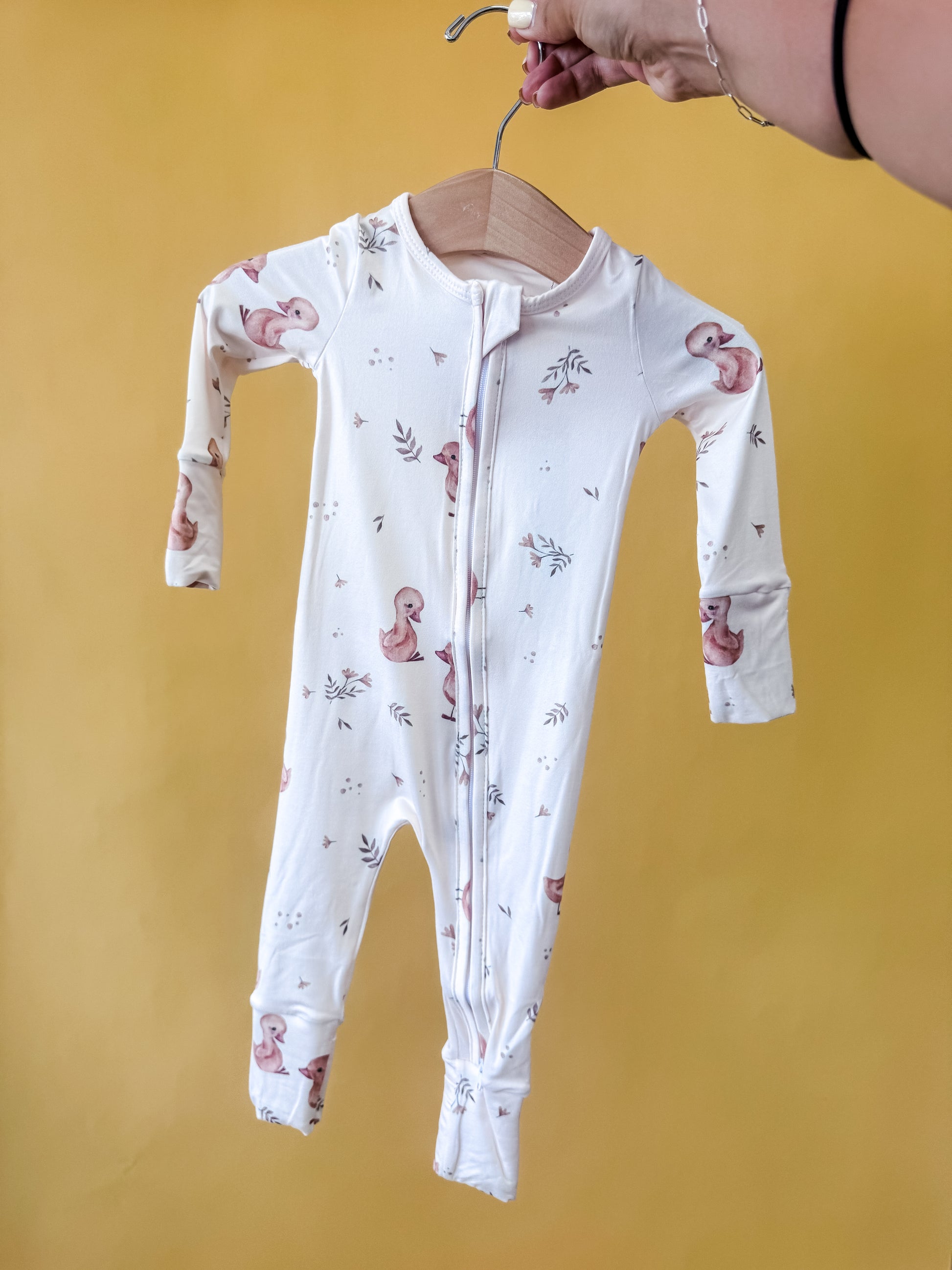 A hand holds a wooden hanger with the Tiny Tot Bundles Morning Waddle Long Zippy, a white baby onesie featuring a zipper and pastel duck and leaf patterns, against a yellow background.