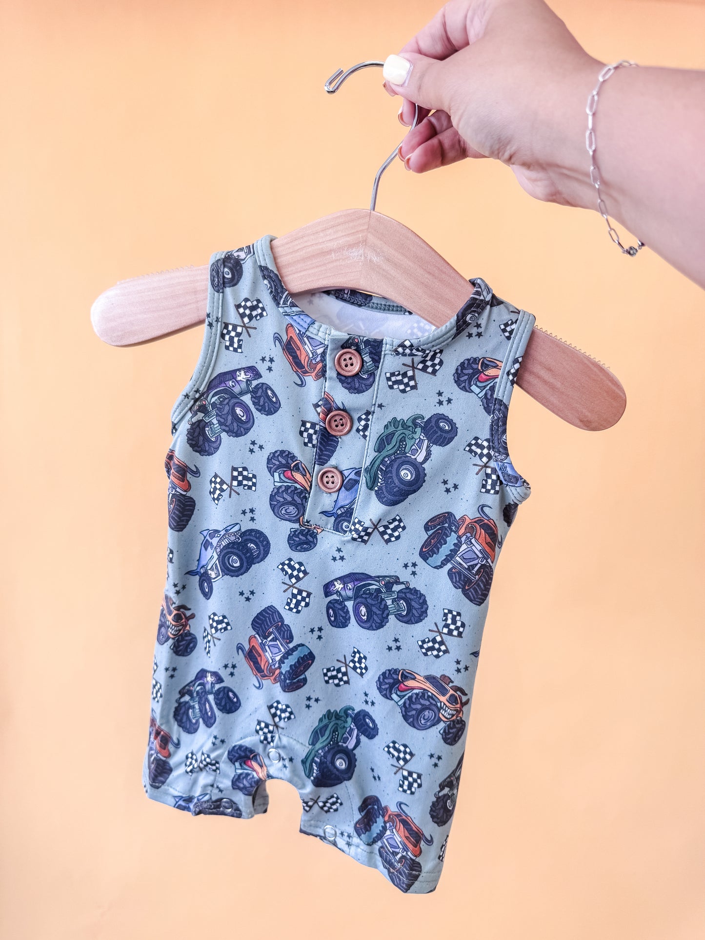 A hand holds a wooden hanger displaying the Tiny Tot Bundles Monster Mash Tank Romper—a sleeveless baby romper featuring colorful monster trucks and checkered flags—set against a peach background.