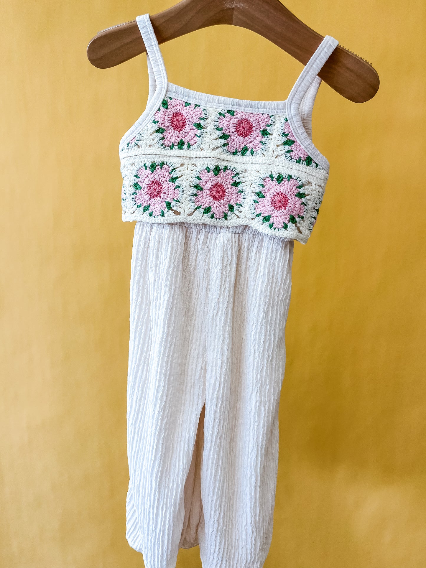 The Petal Patch Jumpsuit by Tiny Tot Bundles, featuring a crocheted top adorned with pink and green flower patterns and textured cream-colored pants, hangs on a wooden hanger against a yellow background.