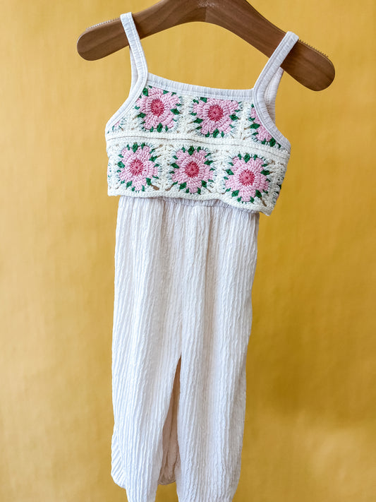 The Petal Patch Jumpsuit by Tiny Tot Bundles, featuring a crocheted top adorned with pink and green flower patterns and textured cream-colored pants, hangs on a wooden hanger against a yellow background.