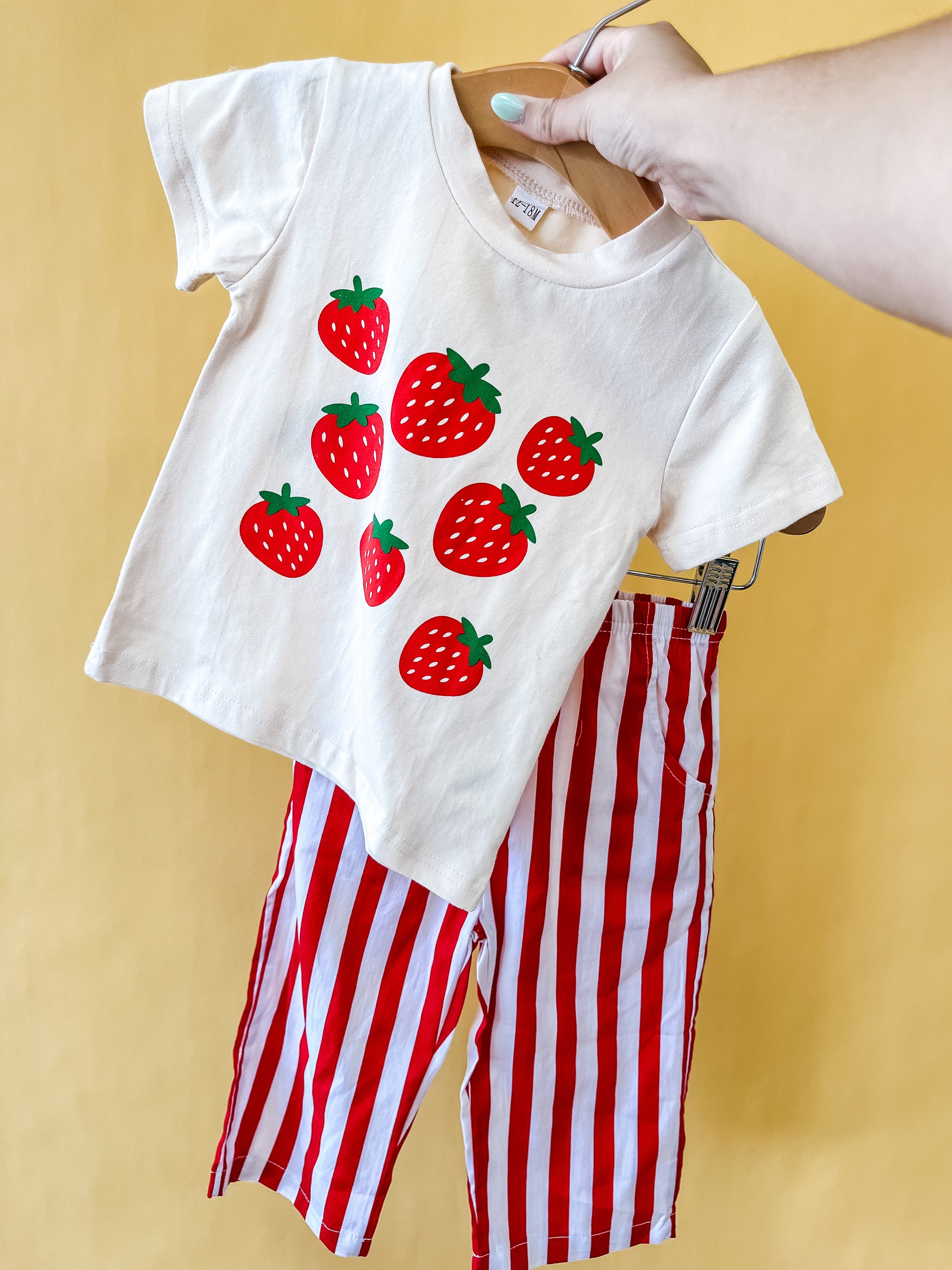 A hand holds the Tiny Tot Bundles Strawberry Patch Set, featuring a children's white T-shirt adorned with red strawberries and matching red-and-white vertically striped pants, set against a yellow background.