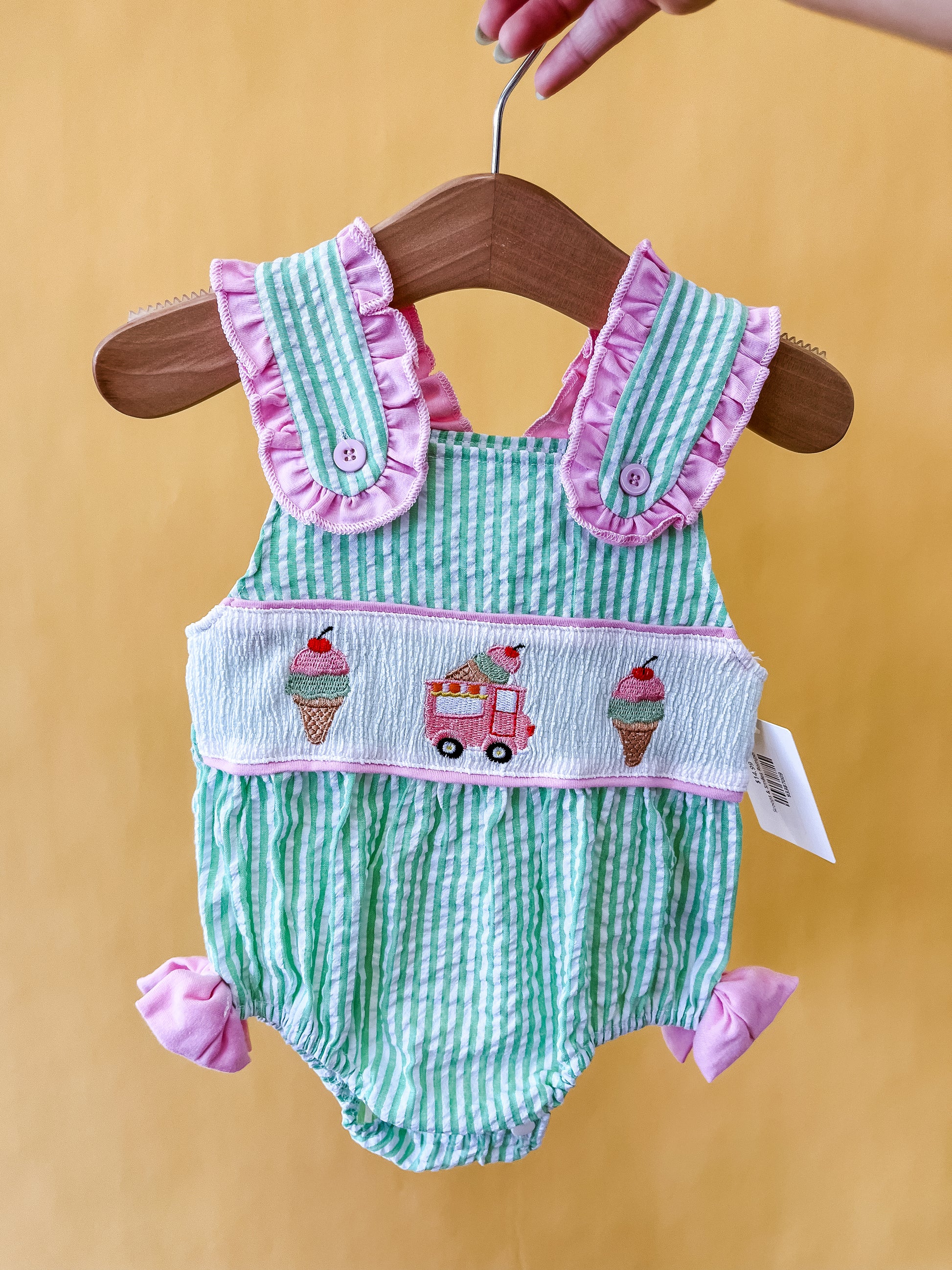 The Tiny Tot Bundles "Scoops & Smiles Romper" features green and white stripes, pink ruffle trim, playful ice cream cone and truck embroidery, and pink bows at the legs, displayed on a wooden hanger against a yellow background.
