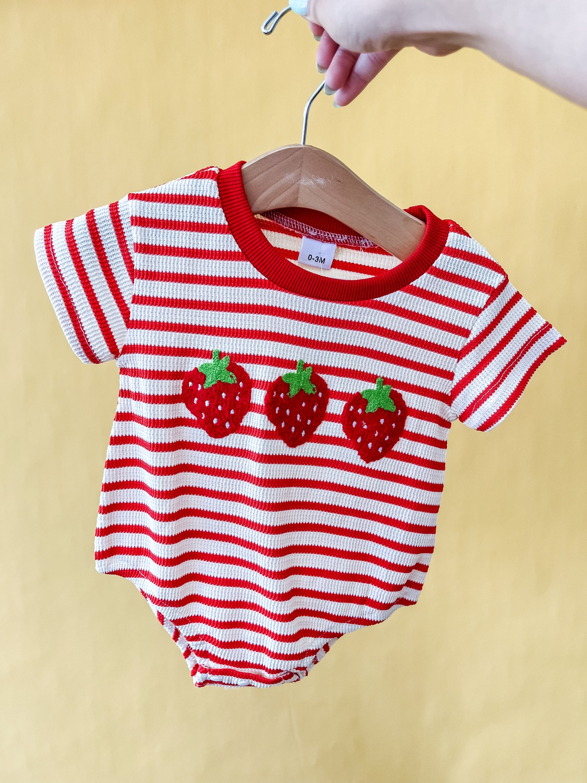 A hand holds the Tiny Tot Bundles "Sweet as Strawberry Bubble" baby romper, featuring red and white stripes and three strawberry appliqués on the front, displayed on a wooden hanger against a yellow background.