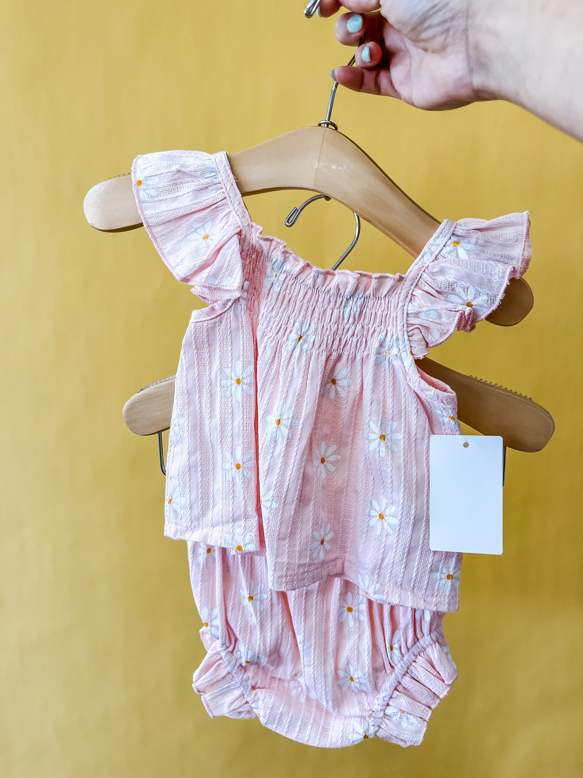 A hand holds a hanger with the Dainty Floral Infant Set by Tiny Tot Bundles, featuring ruffled sleeves and charming white daisy patterns on pink fabric, displayed against a solid yellow background.