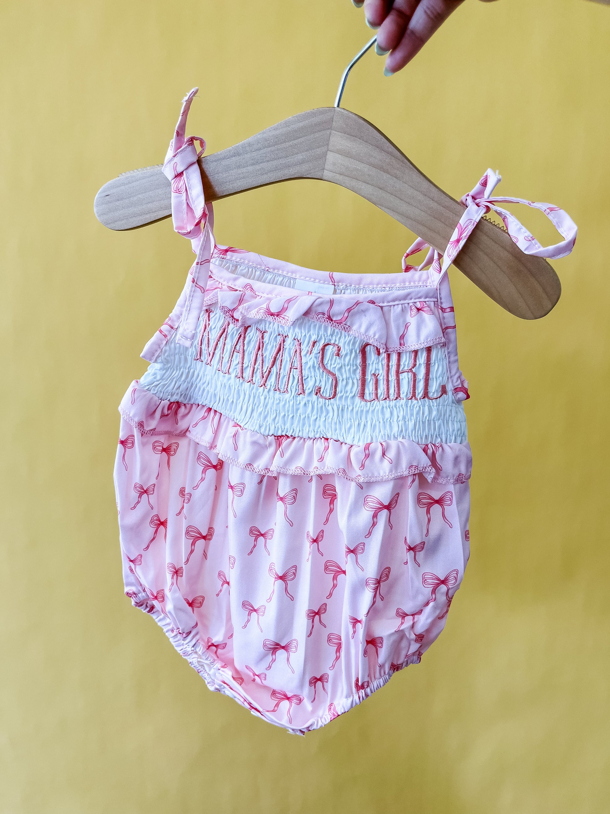 The Mama's Girl Bow Romper by Tiny Tot Bundles, featuring adorable bows and "MAMA'S GIRL" embroidered on the chest, is displayed on a wooden hanger against a yellow background.