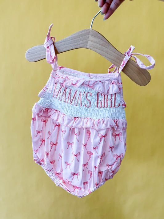 The Mama's Girl Bow Romper by Tiny Tot Bundles, featuring adorable bows and "MAMA'S GIRL" embroidered on the chest, is displayed on a wooden hanger against a yellow background.