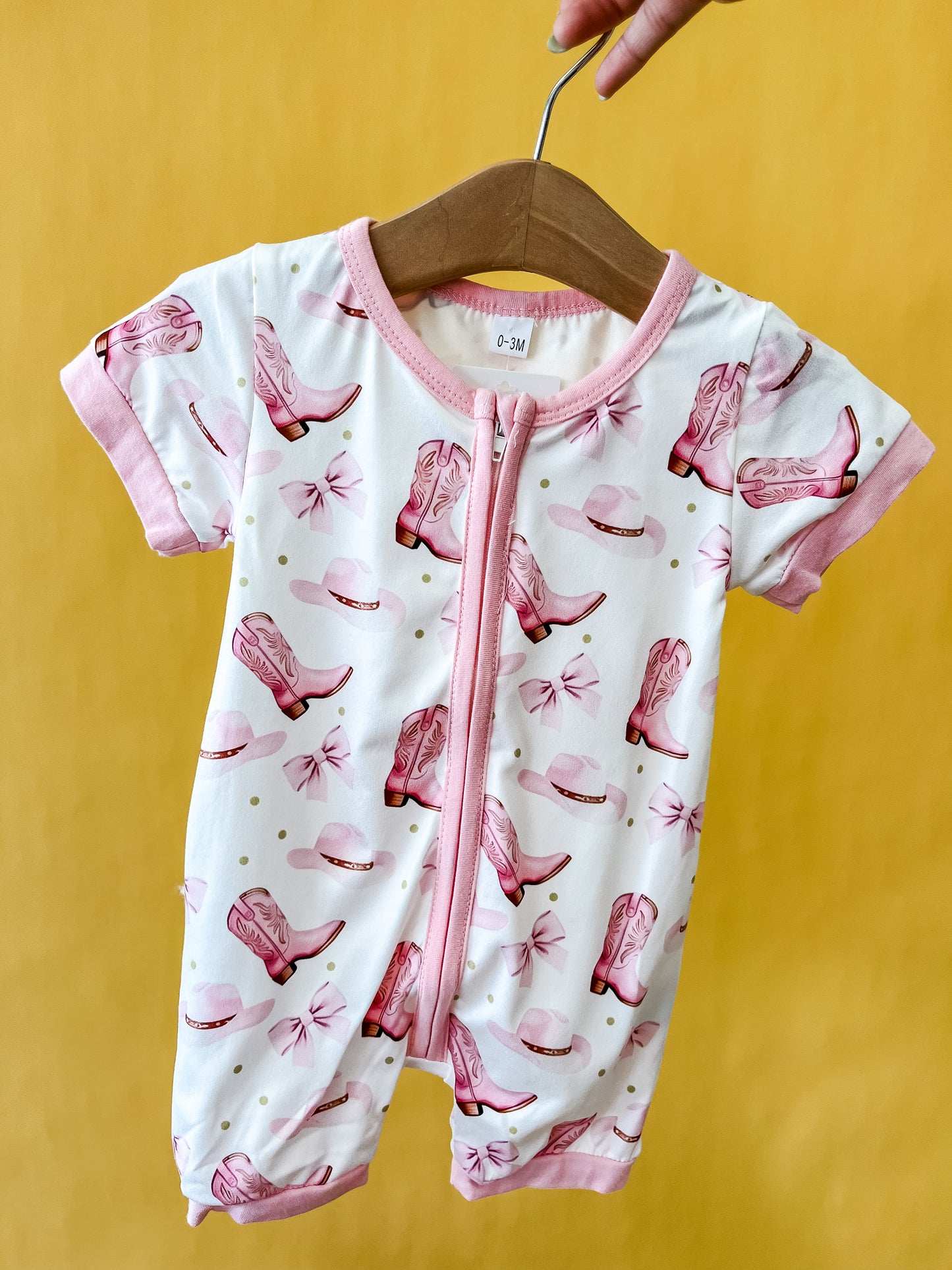 The Pink Boots & Bows Romper by Tiny Tot Bundles is a short-sleeved baby romper with pink trim, featuring a white background decorated with a pink cowboy hat and boots pattern. Displayed on a hanger held by hand against a solid yellow background.