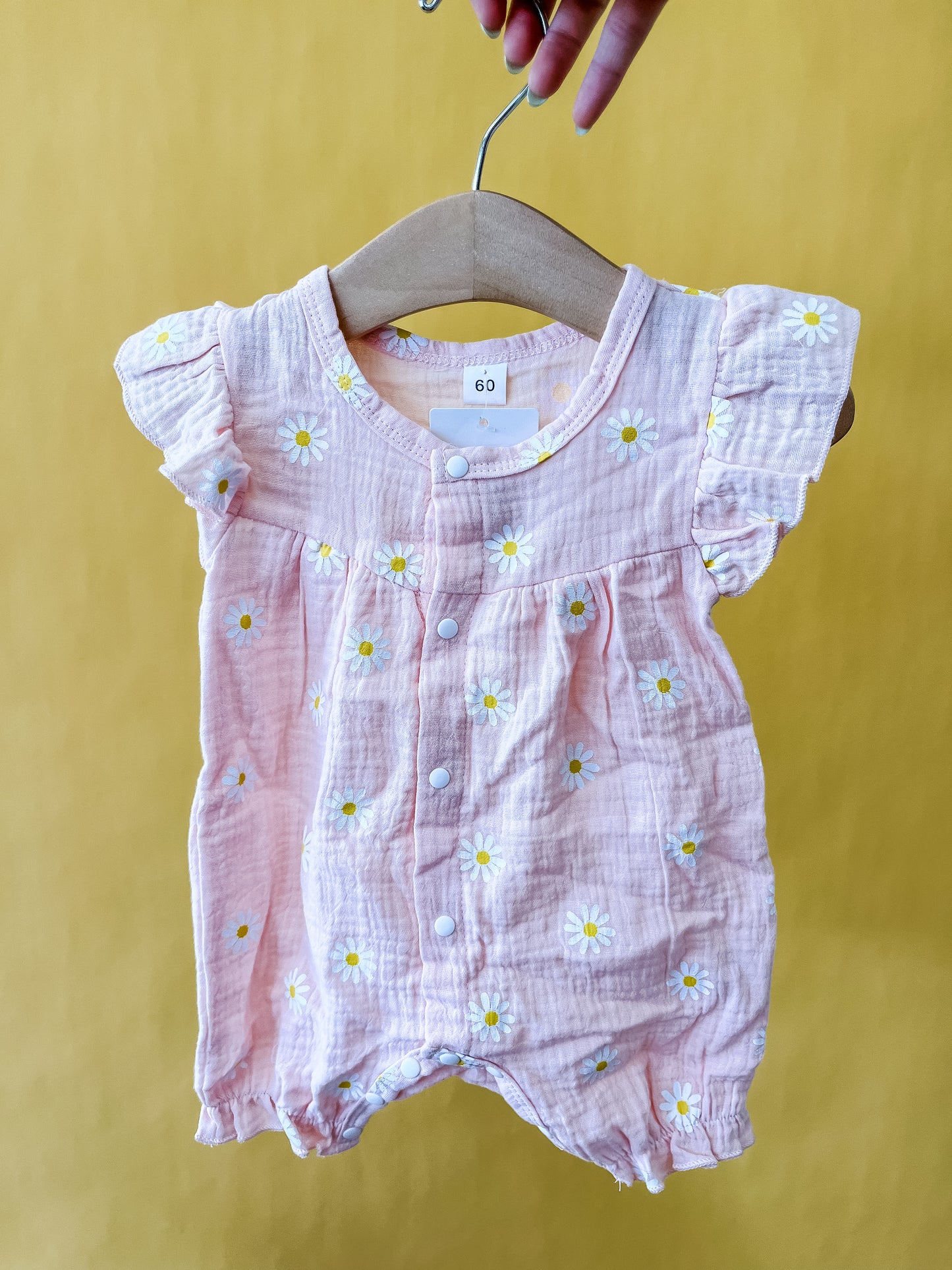 The Daisy Dreams Romper by Tiny Tot Bundles, featuring white and yellow daisy prints, ruffled sleeves, and front buttons in a soft pink hue, is displayed on a wooden hanger against a yellow background.